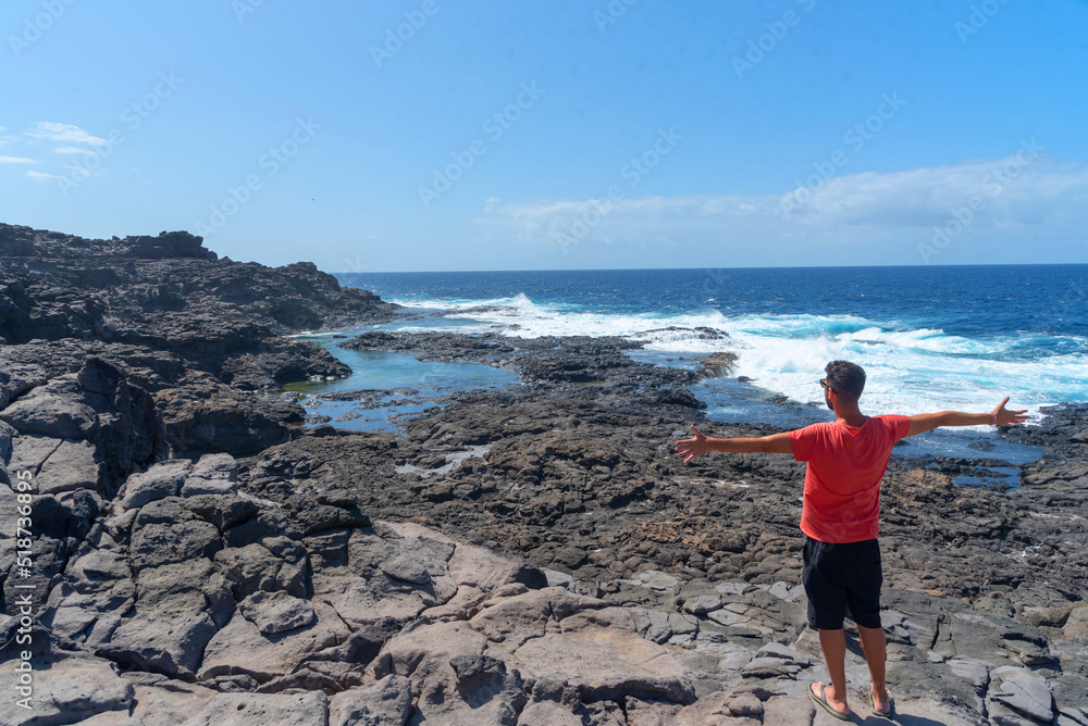 Hombre moreno de espaldas con camiseta roja y los brazos abiertos en la costa de Lanzarote disfrutando de unas piscinas naturales con el mar de color turquesa en Islas Canarias.