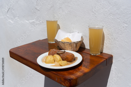 Plato de patatas con salsa y una croqueta de jamón y queso sobre un plato blanco acompañado por dos cervezas frescas y una cestita de mimbre con pan, sobre una mesa de madera con fondo blanco photo