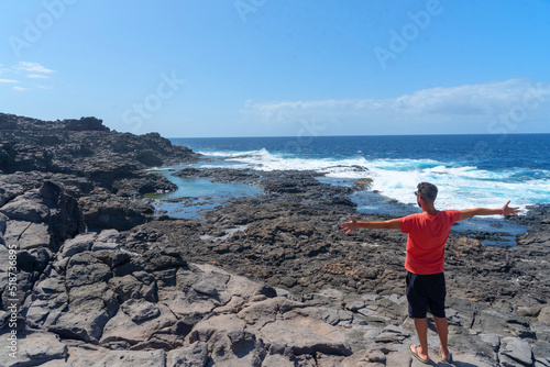 Hombre moreno de espaldas con camiseta roja y los brazos abiertos en la costa de Lanzarote disfrutando de unas piscinas naturales con el mar de color turquesa en Islas Canarias.
