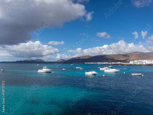 Punta Mujeres con su arquitectura tradicional de casas blancas visto desde el mar turquesa de Lanzarote con alg  n barco y grandes volcanes inactivos detr  s en las Islas Canarias