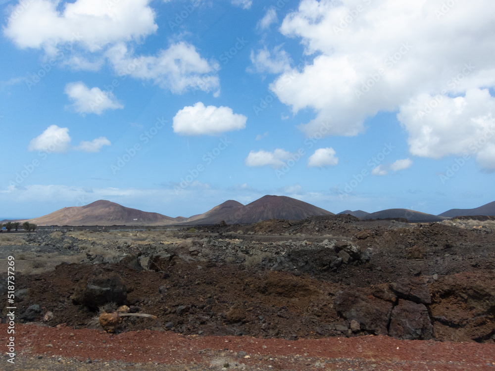 Paisaje volcánico con grandes volcanes inactivos al fondo y terreno con rocas y tierra negra durante un día soleado con alguna nube en Lanzarote, Islas Canarias.