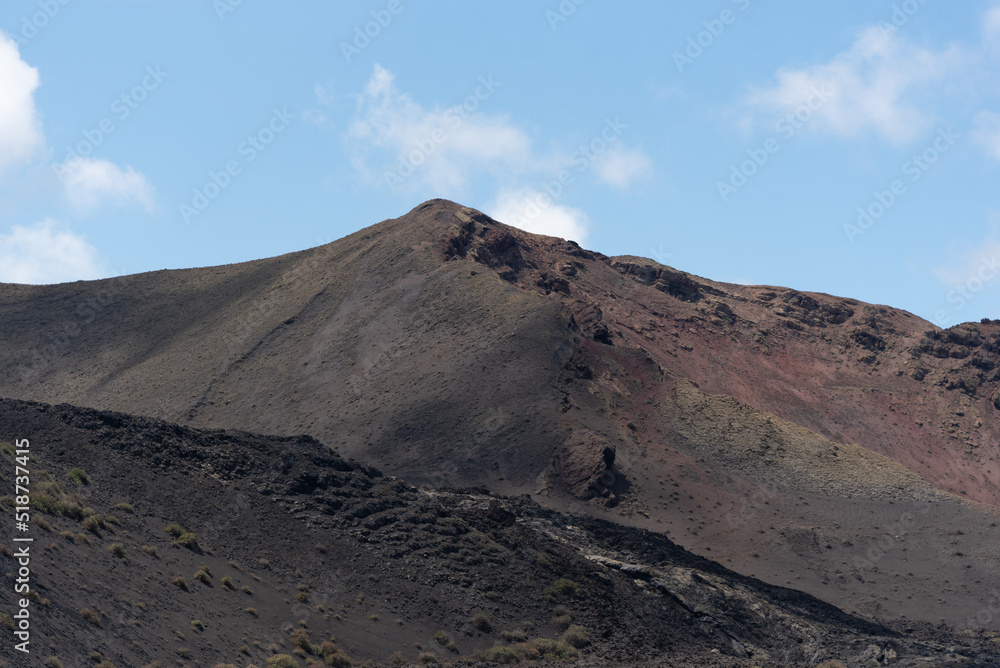 Paisaje volcánico y desértico con tierra negra y rocas oscuras y cos volcanes inactivos durante un día de verano en Lanzarote, Islas Canarias. Recursos turísticos y naturales.