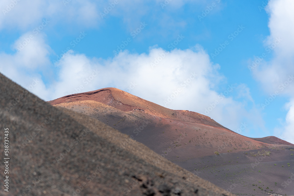 Detalle de un paisaje desértico y volcánico de tierras en tonos marrones y naranjas con un gran volcán inactivo al fondo durante un día soleado de verano en Lanzarote, Islas Canarias.