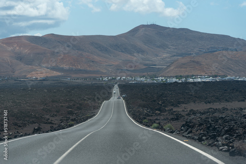 Vista panorámica de un paisaje volcánico con una carretera en medio con dos grandes volcanes inactivos al fondo durante un día soleado con el cielo azul despejado en Lanzarote, Islas Canarias. 