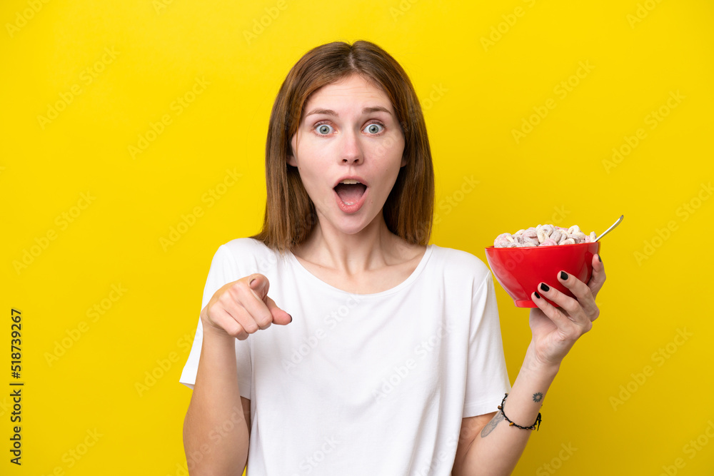 Young English woman holding cup of coffee isolated on yellow background surprised and pointing front Young English woman holding cup of coffee isolated on yellow background surprised and pointing front