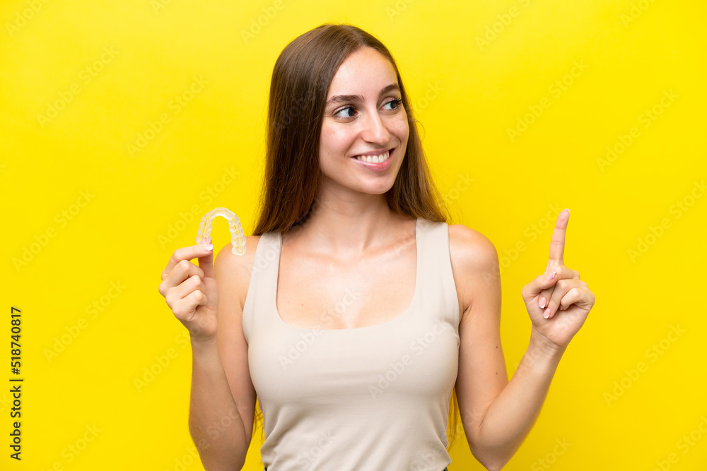 Young caucasian woman holding invisible braces isolated on yellow background intending to realizes the solution while lifting a finger up Young caucasian woman holding invisible braces isolated on yellow background intending to realizes the solution while lifting a finger up