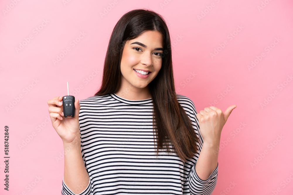 Young Brazilian woman holding car keys isolated on pink background pointing to the side to present a product Young Brazilian woman holding car keys isolated on pink background pointing to the side to present a product