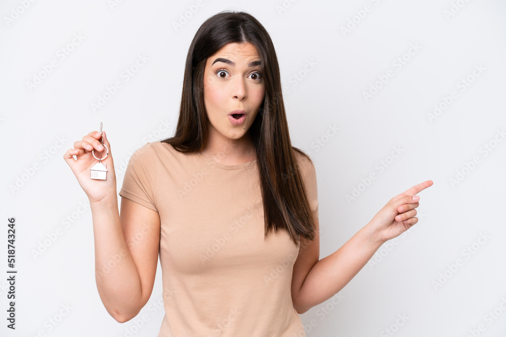 Young Brazilian woman holding home keys isolated on white background surprised and pointing side Young Brazilian woman holding home keys isolated on white background surprised and pointing side