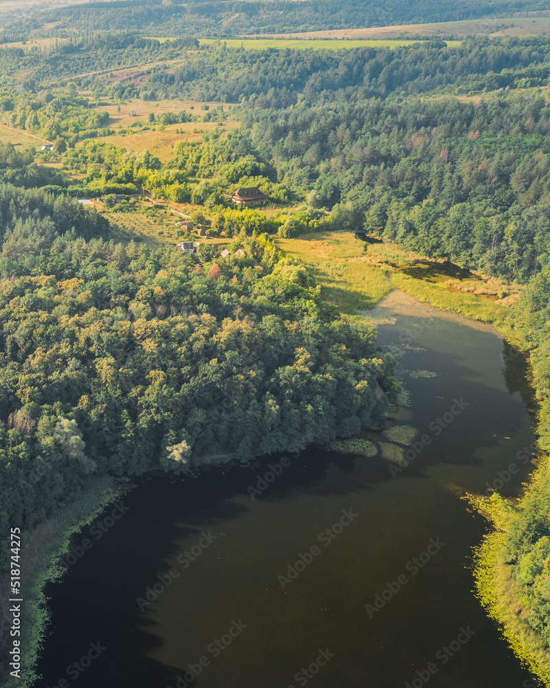 House on plot on the shore of a large lake among green forests with trees - aerial drone shot. Ecological settlement among the forest on the shore of a wild lake: wild life in the forests of Ukraine.