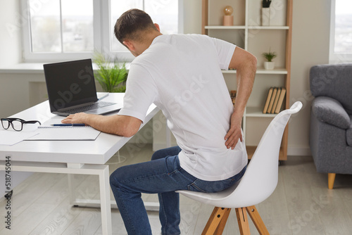 Man experiences severe back pain caused by prolonged and incorrect sitting at workplace. Young man sitting with his back to camera holds his lower back, feeling exhausted from sedentary lifestyle. photo