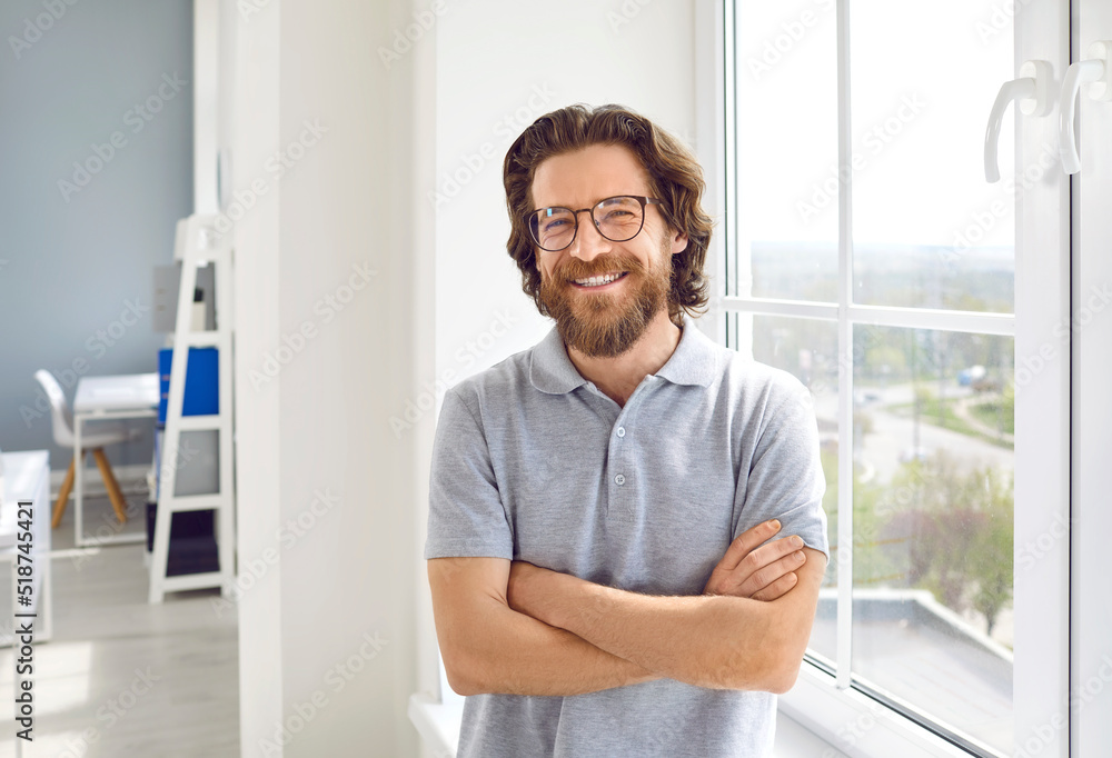Portrait of a happy successful businessman. Cheerful smiling handsome bearded young Caucasian business man in a casual grey polo shirt and glasses standing with his arms crossed by the office window