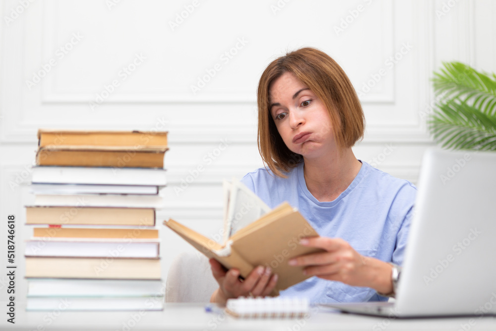 Tired and bored female student reading book and preparing for exams at university. Stack of books and laptop on the desk. Online education and learning from home concept Tired and bored female student reading book and preparing for exams at university. Stack of books and laptop on the desk. Online education and learning from home concept