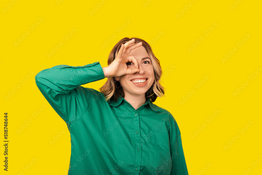 A photo of a cheerful young woman that i showing the ok sign holding it near her eye smiling at the camera A photo of a cheerful young woman that i showing the ok sign holding it near her eye smiling at the camera