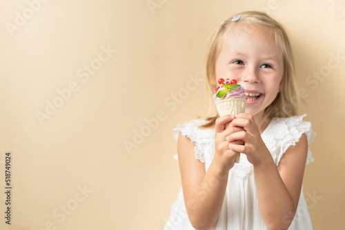 Beautiful little caucasian girl with blonde hair in a white dress enjoying berry flavored waffle cone ice cream on the beige background