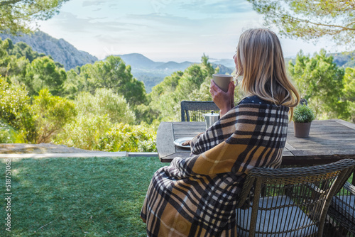 Woman drinking coffee on the terrace on a sunny day. Female enjoys nature and fresh air in a plaid. Autumn style.
