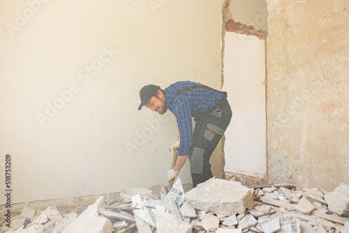Obraz na plátne A man in a work suit with a baseball cap cleans up in an apartment under renovation, picks up debris from the floor left after demolishing walls with a shovel