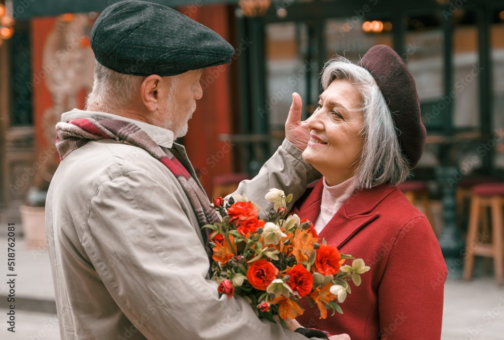 Elderly senior love couple. Old retired man woman together on romantic date.Aged husband wife walking on city street with flowers.Stylish elder hugging people pensioner in red coat.Happy family years