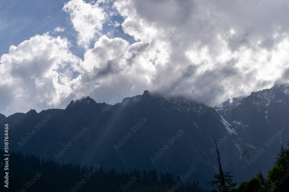 Mountains covered in dense clouds and sunlight is coming through clouds. Dense cedar forest and green meadow creates a beautiful landscape on a rainy day in Manali, Himachal Pradesh in India.