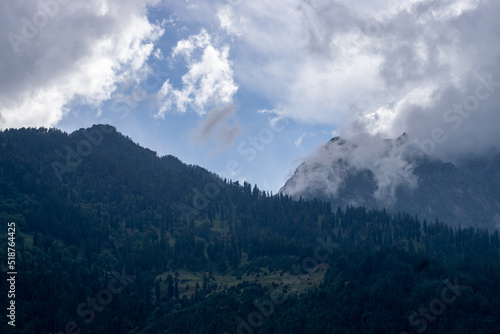 Mountains covered in dense clouds and sunlight is coming through clouds. Dense cedar forest and green meadow creates a beautiful landscape on a rainy day in Manali, Himachal Pradesh in India.