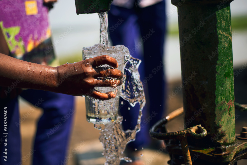 Children having fresh water from the tube well in a rural village to hydrate the body in the hot summer. Hand holding glass and splash of clean water from the tube well.