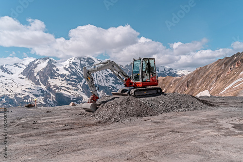 red excavator with shovel standing on hill with stones, mountains in background
