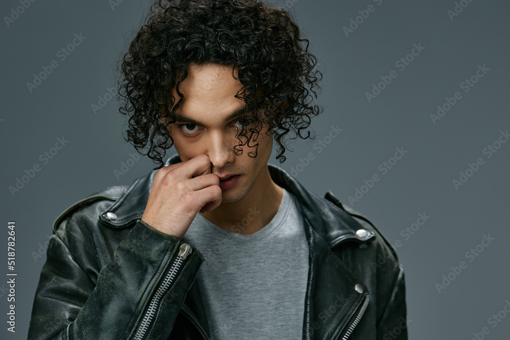 Obraz premium Sexy pretty stylish tanned curly man leather jacket looks at camera posing isolated on over gray studio background. Cool fashion offer. Huge Seasonal Sale New Collection concept. Copy space for ad