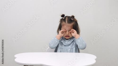 Cute capricious girl is crying while sitting at the table on a white background. Education.