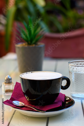 Fototapeta Naklejka Na Ścianę i Meble -  Cup of cappuccino coffee in black cup, spoon on saucer and glass of water on a wooden white table at the cafe. Vertical view