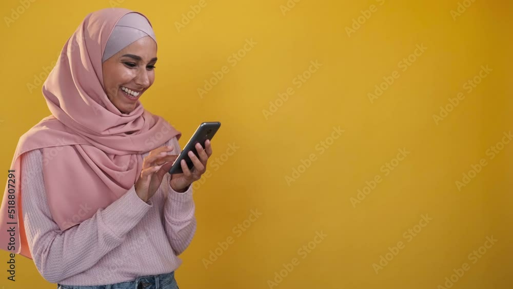 Gadget user. Chatting online. Internet communication. Cheerful woman in hijab texting message on phone isolated on orange copy space background. Gadget user. Chatting online. Internet communication. Cheerful woman in hijab texting message on phone isolated on orange copy space background.