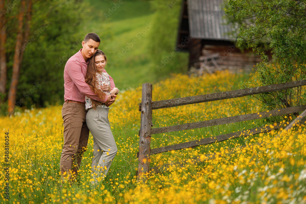 Young loving couple gently hugging on the wood house background, a lot of yellow flowers are around on the field. Enjoying time together. Young loving couple gently hugging on the wood house background, a lot of yellow flowers are around on the field. Enjoying time together.