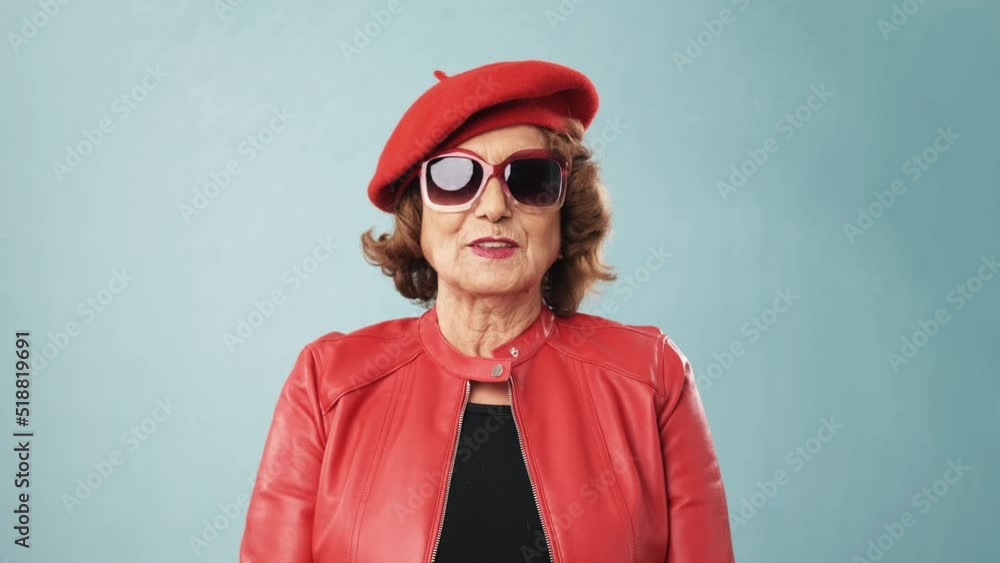 Stylish senior woman in sunglasses wearing a red jacket and a hat while smiling Stylish senior woman in sunglasses wearing a red jacket and a hat while smiling