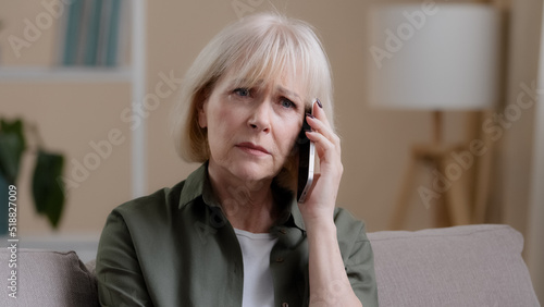 Caucasian old woman mature middle-aged 50s offended lady aged businesswoman sitting on sofa at home listening to voice on phone talking smartphone receiving bad news sadness worry upset losing failure