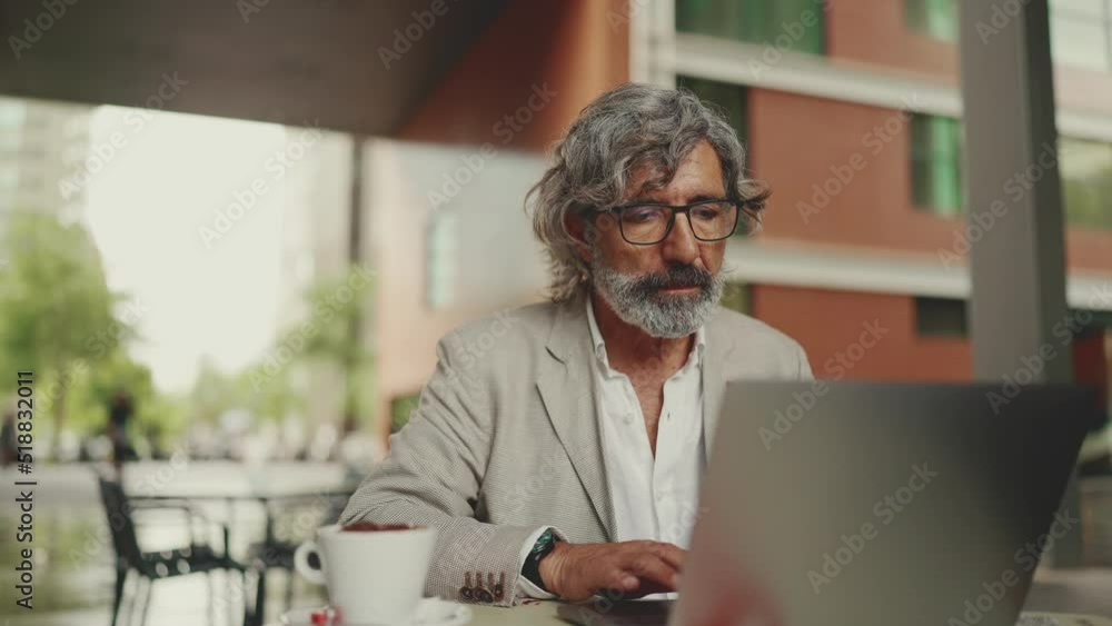 Mature businessman with beard in eyeglasses sits in cafe. Middle aged manager successful man working on laptop pc computer sit at cafe outdoor. Takes off glasses and rubs eyes Mature businessman with beard in eyeglasses sits in cafe. Middle aged manager successful man working on laptop pc computer sit at cafe outdoor. Takes off glasses and rubs eyes