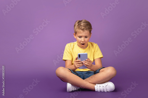 Little shocked kid boy preteen looking to the cell phone. Surprised child using mobile phone sitting on the floor on purple background. Studio shot.