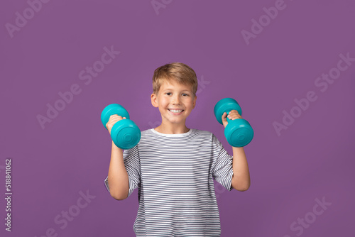 Happy smiling blond boy using fitness equipment, holding green dumbbell, building arm muscle, looking at camera staying on purple background. Studio shot.