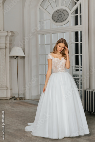 The beautiful woman posing in a wedding dress. Young beautiful happy bride wearing white wedding dress and posing in bright empty interior. Wedding fashion. Expensive Lace bust.