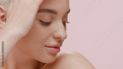 Good-looking young slim Caucasian woman touches her half-turned head smiling on pale pink background | Face hydration concept