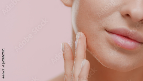 Extreme close-up shot of attractive young slim white-skinned woman strokes her jawline smiling for the camera on pale pink background | Anti-aging concept