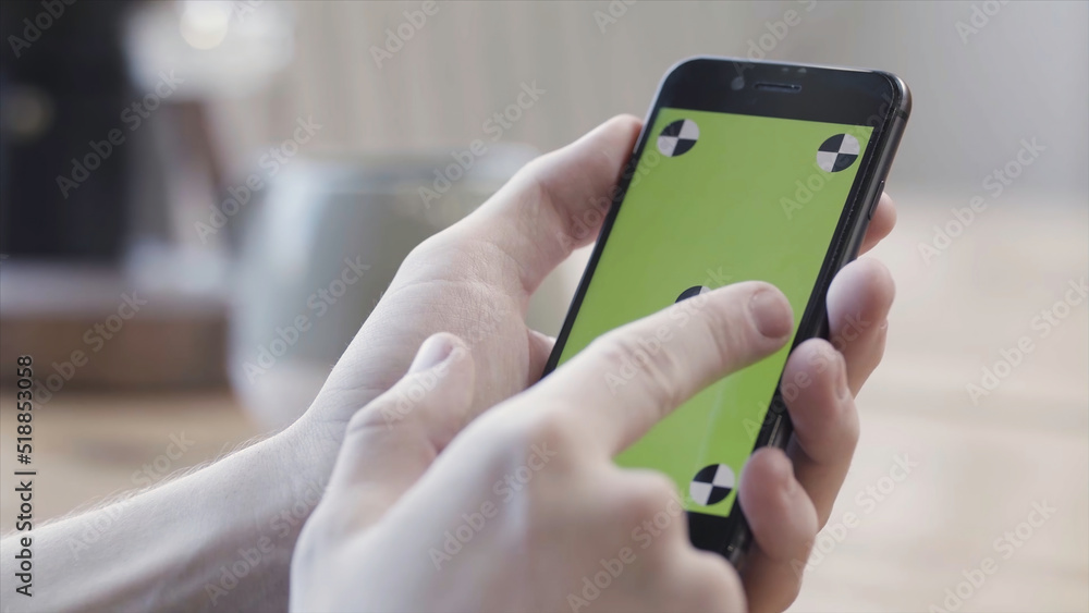 Close up of hands using smartphone with green screen, sliding and tapping on blurred kitchen table background. Stock footage. Man hands holding mobile phone with chroma key.