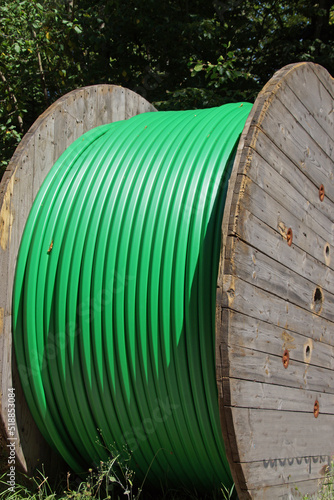 Mannshohe Kabeltrommel aus Holz. Aufgewickelt ein dickes Kunststoff-Kabel. Es ist deutlich grün. Die Trommel steht am Straßenrand an einer grünen Hecke. Baustelle ist nebenan. 