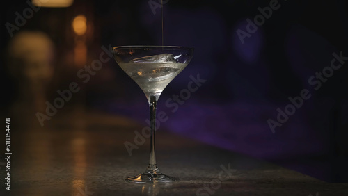 Close up of pouring alcohol into the martini glass with ice cube. Stock footage. Thick alcoholic beverage is being poured into transparent glass on dark background.