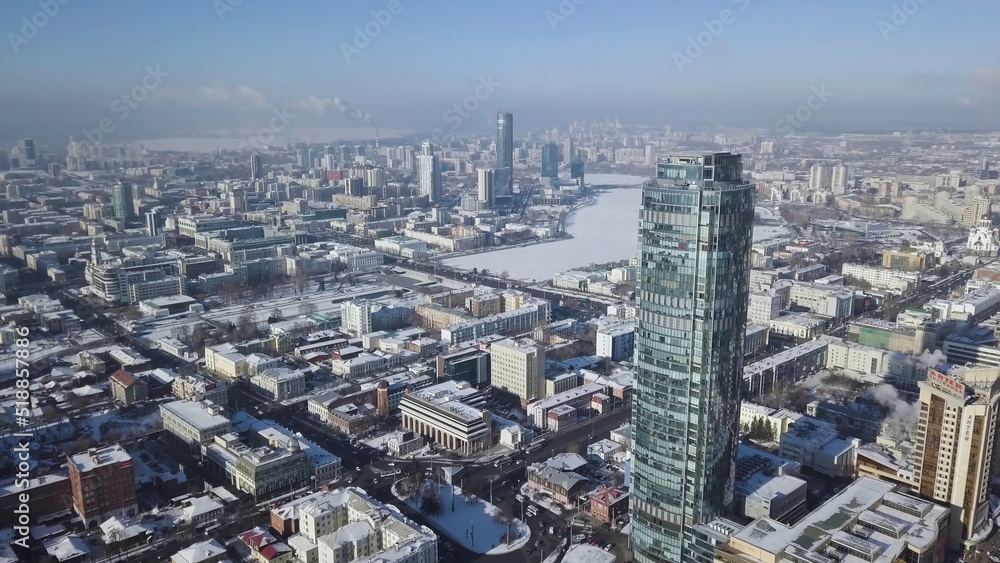 Fototapeta premium Top view of the amazing glass tower or the business center in the background of a winter city. Aerial view of skyscraper is in the middle of the city in winter, blue sky sky and snowy roofs of