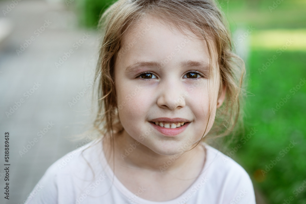 Child's portrait girl in light T-shirt close-up against background green grass smiling sweetly and looking at the camera. Caucasian little girl Child's portrait girl in light T-shirt close-up against background green grass smiling sweetly and looking at the camera. Caucasian little girl