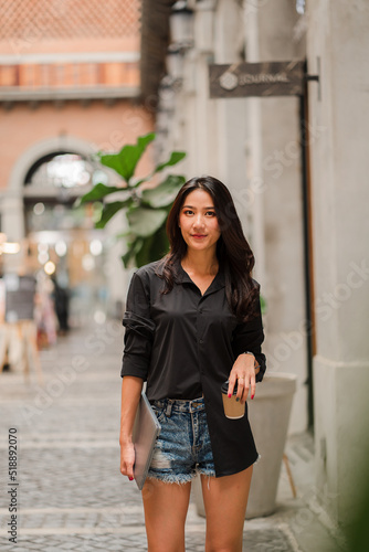 Beautiful Asian woman holding a cup of coffee with a laptop. and standing in front of the office building to go to work in the morning © ArLawKa