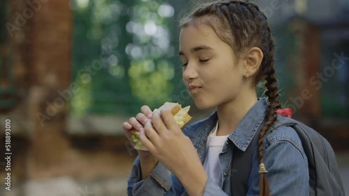 A little schoolgirl is eating a sandwich standing during the break on the schoolyard. The child stands and eats bread and cheese on the street. Food nutrition and education concept