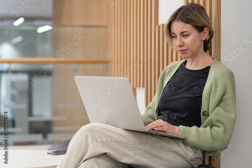 Serious woman holding smartphone discussing project details while working online on laptop in coworking space, talking with client via cellphone, businesswoman with mobile phone looking at pc screen