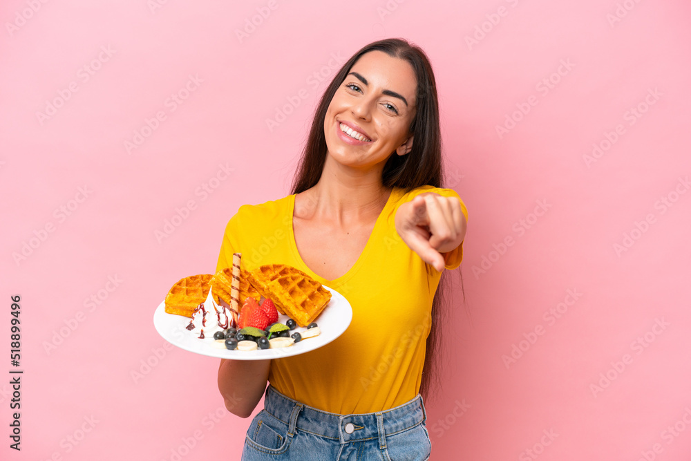 Young caucasian woman holding waffles isolated on pink background pointing front with happy expression Young caucasian woman holding waffles isolated on pink background pointing front with happy expression