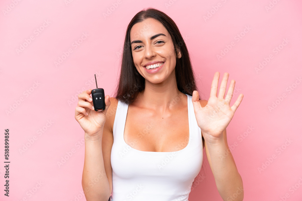 Young caucasian woman holding car keys isolated on pink background saluting with hand with happy expression Young caucasian woman holding car keys isolated on pink background saluting with hand with happy expression