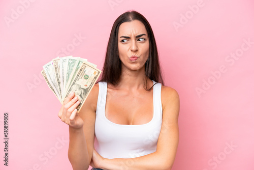 Young caucasian woman taking a lot of money isolated on pink background with sad expression