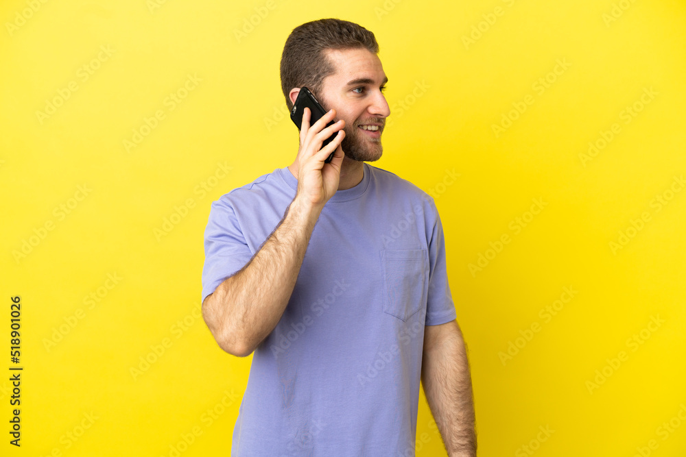 Handsome blonde man over isolated yellow background keeping a conversation with the mobile phone Handsome blonde man over isolated yellow background keeping a conversation with the mobile phone